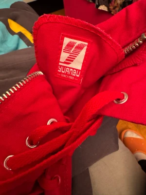 Röda höga sneakers från Yuanbu - Snygga röda sneakers från Yuanbu med högt skaft, vita gummisulor och klassisk snörning. Skorna har dragkedja på sidan och metallöljetter för snörena. Tungan har en tydlig logga och materialet är slitstark textil.