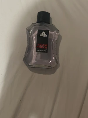 Adidas Team Force Eau de Toilette - Adidas Team Force Eau de Toilette i en transparent, vågformad glasflaska med svart skruvkork. Flaskan har en svart etikett med Adidas-loggan och röd text. Doften är sportig och energisk, perfekt för dig som gillar fräscha och maskulina parfymer.
