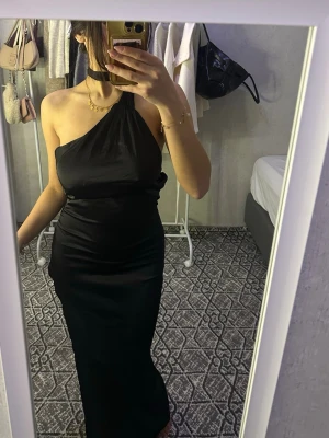 Svart oneshoulder långklänning - Snygg svart långklänning med oneshoulder-design och hög slits. Klänningen har öppen rygg och är figurnära med elegant fall. Perfekt för dig som vill sticka ut med en stilren look.