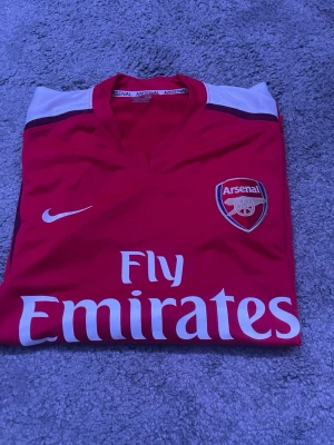 Arsenal Nike matchtröja röd/vit - Säljer en klassisk Arsenal matchtröja från Nike i rött med vita ärmar och svarta detaljer. Tröjan har Arsenal-logga på bröstet, Nike-logga och Fly Emirates-tryck framtill. Tillverkad i lätt och ventilerande material, perfekt för fotbollsträning eller support.
