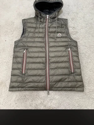 Grå dunväst med huva från Moncler - Stilren grå dunväst från Moncler med huva och dragsko. Västen har två fickor med dragkedja och röda/vita detaljer samt Moncler-logga på bröstet. Perfekt för lager-på-lager och streetstyle.
