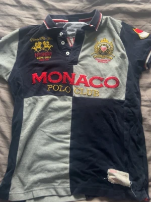 Monaco Polo Club pikétröja - Snygg pikétröja från Monaco Polo Club med broderade detaljer och loggor i rött, gult och vitt. Tröjan har blockmönster i marinblått och grått, klassisk krage med knappar och korta ärmar. Perfekt för en sportig och preppy look.