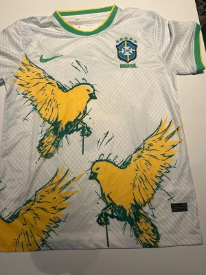 Vit Brasilien fotbollströja Nike - Vit Brasilien fotbollströja från Nike med gröna och gula detaljer. Framsidan har stora gula papegojor i grafisk stil och Brasilien-logga på bröstet. Kragen och ärmsluten har gröna och gula ränder. Perfekt för dig som älskar fotboll och vill sticka ut.