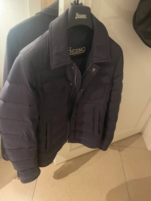 Herno jacka  - Snygg mörkblå pufferjacka från Herno med klassisk krage, flera fickor med lock och både dragkedja och knappar framtill. Jackan har quiltad design och långärmade ärmar, perfekt för kalla dagar. Kommer med original galge och Herno-väska. Har även kvitto och den är köpt på Farfetch tidigare i år men knappast använd den har dock en mindre defekt på ena armen inget som syns men kom privat så kan jag visa. Nypris runt 10.000 kr.