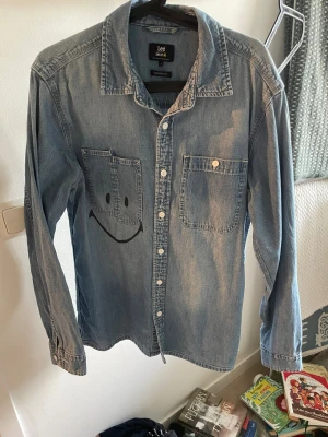 Lee jeansskjorta med smiley-tryck - Snygg ljusblå jeansskjorta från Lee med två bröstfickor och ett stort svart smiley-tryck på ena fickan. Skjortan har klassisk krage, knäppning framtill och långa ärmar. Perfekt för dig som gillar en chill och lekfull stil.
