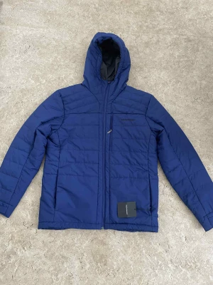 Blå pufferjacka Peak Performance XL - Blå pufferjacka från Peak Performance i storlek XL. Jackan har huva, hel dragkedja framtill och en bröstficka med dragkedja. Quiltad design och diskret logga på bröstet. Perfekt för kalla dagar och riktigt snygg till streetwear.