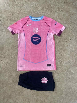 Barça Lamine Yamal #10 rosa matchtröja - Säljer en FC Barcelona matchtröja med Lamine Yamal och nummer 10 på ryggen. Tröjan är rosa med blå detaljer, har korta ärmar och är tillverkad i lätt Dri-Fit material. Fram finns Spotify-logga och klubbmärke, bak UNHCR-tryck. Shorts i mörkblått med klubbmärke ingår.