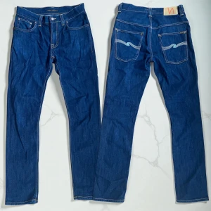 Mörkblå jeans från Nudie Jeans Co - Snygga mörkblå jeans från Nudie Jeans Co med klassisk femficksdesign och kontrastsömmar. Modellen har raka ben och ikonisk broderad detalj på bakfickorna. Jeansen är tillverkade i slitstarkt bomullsjeansmaterial och har knappgylf.