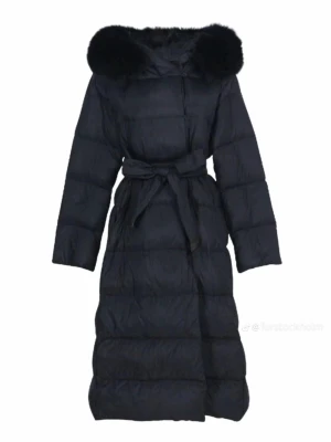 Svart lång dunjacka med pälskrage från FURSWEDEN - Lång svart dunjacka med fluffig pälskrage och bälte i midjan. Jackan har quiltad design och långa ärmar, perfekt för kalla dagar. Snygg och stilren modell som ger en cool siluett.