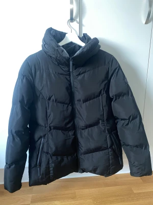 Svart pufferjacka med huva - Säljer en svart pufferjacka med hög krage som går att göra till en huva. Jackan har dragkedja framtill och två snedställda fickor med dragkedja. Den är vadderad och har en clean, stilren look som passar perfekt när det är kallt ute. Har lite sminkfläckar, har dock fått bort de mesta. 