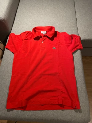 Röd pikétröja från Lacoste, M - Snygg röd pikétröja från Lacoste i klassisk passform. Tröjan har korta ärmar, krage med två knappar och den ikoniska gröna krokodilloggan broderad på bröstet. Materialet är mjuk bomull som andas och känns skönt mot huden. Perfekt för dig som gillar stilren och sportig look.