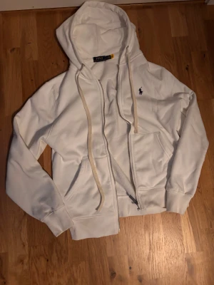 Vit zip hoodie från Polo Ralph Lauren - Säljer en fräsch vit hoodie från Polo Ralph Lauren med klassisk blå logga på bröstet. Tröjan har huva med snörning, dragkedja framtill och två stora fickor. Perfekt för chill dagar och enkel att matcha med jeans eller joggers. Säljer pga att den är för liten i storleken, endast använd ett fåtal gånger. Storlek M men sitter som S
