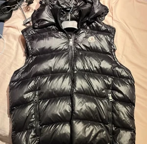 Svart dunväst från Moncler - Svart dunväst från Moncler med glansig finish och klassisk logga på bröstet. Västen har huva, två framfickor med dragkedja och quiltad design. Insidan har ett unikt serietidningsmärke. Perfekt för att hålla värmen och se stylish ut.
