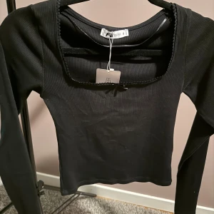 Svart ribbad topp från new yorker  - Svart långärmad topp från new Yorker  med fyrkantig ringning och dekorativ kant. Ribbad struktur i stretchigt material som sitter snyggt på kroppen. Perfekt att styla med jeans eller kjol för en clean look.