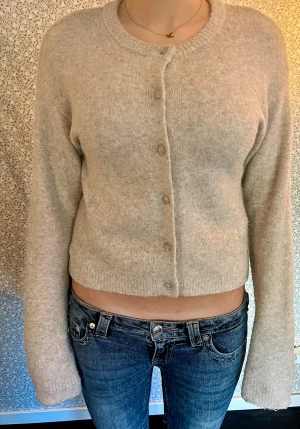 Beige kofta med knappar - Supermysig beige kofta med croppad passform och rund hals. Tröjan har långa ärmar och stängs med diskreta knappar framtill. Materialet är mjukt och stickat.