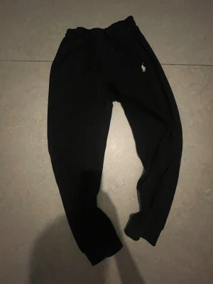 Svarta mjukisbyxor från Polo Ralph Lauren - Svarta mjukisbyxor från Polo Ralph Lauren med klassisk logga broderad på höften. Byxorna har elastisk midja med snörning och ribbade muddar vid bensluten. Perfekta för chill eller träning, riktigt sköna och stilrena. Passar till 160-165
