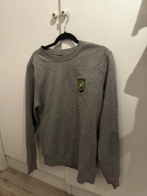 Grå Hammarby sweatshirt från UB - Grå sweatshirt från Clique i storlek S med Hammarby-emblem broderat på bröstet. Tröjan har rund hals, långa ärmar och ribbade muddar. Perfekt för dig som vill visa ditt stöd för Hammarby och gillar enkel, sportig stil.