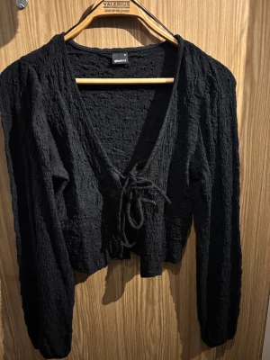 Svart croppad omlottopp från Gina Tricot - Svart croppad omlottopp från Gina Tricot med lång ärm och snygg snörning framtill. Toppen har en cool textur i tyget och djup v-ringning. Perfekt för dig som gillar stilrena och trendiga plagg med lite edge.