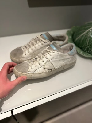 Philippe Modell skor - Säljer ett par snygga Philippe Model sneakers i vitt och grått med ljusblå detalj på hälen och tungan. Skorna är tillverkade i skinn och mocka. Klassisk låg modell med platt sula och ikonisk sköld på sidan. Skorna är rätt använda men absolut i ett brukbart skick och ger för övrigt en rätt okej helheltskänsla enligt mig. Super prisvärt för dessa äkta Philippe model skor ifall du letar efter stil till din garderob! 🤗 Pris är ej hugget i sten så tveka ej på att höra av dig! 😊