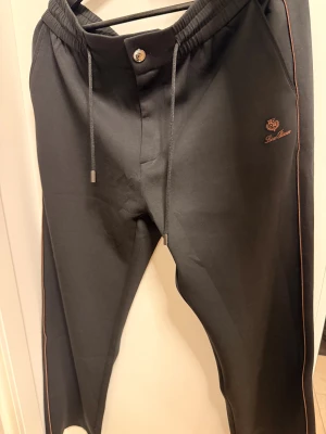 Svarta mjukisbyxor från Loro Piana - Svarta mjukisbyxor från Loro Piana med brunt broderat logomärke på höften och bruna detaljer längs sidorna. Byxorna har elastisk midja med snörning, knapp framtill och bakficka med dragkedja. Sköna och stilrena, perfekta för en chill look.