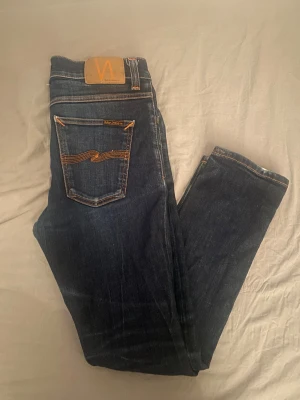 Mörkblå jeans från Nudie Jeans - Säljer ett par mörkblå jeans från Nudie Jeans med klassisk orange söm och snygg broderad bakficka. Jeansen har en slim passform och är tillverkade i slitstarkt denimtyg av bomull. Perfekta för dig som gillar en stilren och modern look. Strl 31 30