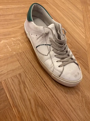 Philippe Model vita sneakers med gröna detaljer - Säljer ett par vita Philippe Model sneakers i skinn med grå snörning och gröna detaljer på hälen. Skorna har en klassisk låg profil, rund tå och diskret mönster på sidan. Perfekta för dig som gillar stilrena och trendiga sneakers. Kan mötas upp i Stockholms innerstad samt i Uppsala på vardagar. #philippe #philippemodel #maison#östermalm #uppsala 