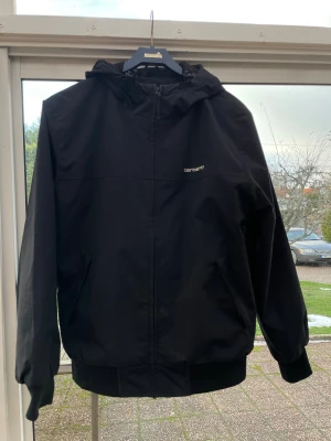 Svart vindjacka från Carhartt WIP - Svart vindjacka från Carhartt Work In Progress med huva och dragkedja. Jackan har två snedställda fickor, ribbade muddar och diskret Carhartt-logga på bröstet. Yttermaterial i polyester och mjukt foder på insidan. Perfekt för blåsiga dagar. Köpt förra året på kennidies knappt använd pga fel storlek väldigt fin 