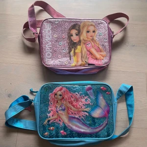 Glittriga top model axelväskor med motiv - Två axelväskor med glittriga framsidor och motiv. Väskorna är från TOPModel har bild på två tjejer och glittriga detaljer, medan den blå från Fantasy Model har en sjöjungfru med rosa hår och havsdjur. Båda har justerbara axelremmar och dragkedja.