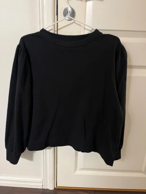 Svart långärmad tröja från 157 - Svart långärmad tröja från 157 med rund hals och puffade ärmar som ger en snygg siluett. Tröjan har en loose passform och är tillverkad i mjuk bomull, perfekt för dig som gillar enkel och stilren look. Passar dig som vill ha något bekvämt och trendigt.
