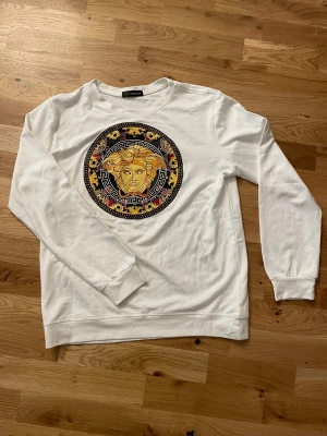 Vit Versace sweatshirt med Medusa-tryck - Vit sweatshirt från Versace med stort, färgstarkt Medusa-tryck i gult, svart och rött på bröstet. Tröjan har rund hals, långa ärmar och ribbade muddar. Tillverkad i mjuk bomull för en skön känsla och snygg passform.