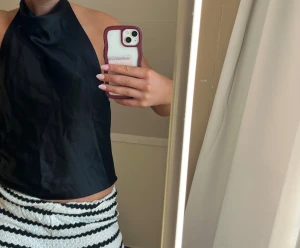 Svart halterneck linnetopp - Säljer en svart halterneck linnetopp med hög hals och ärmlös design. Toppen har en slät, lätt glansig yta och croppad passform som ger en trendig look. Perfekt för att styla med både jeans och kjol.