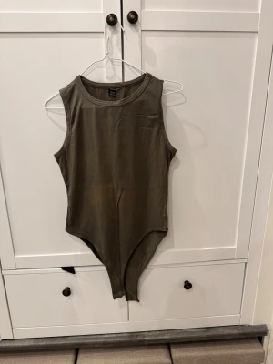 Olivgrön ärmlös bodysuit från SHEIN - Säljer en stilren olivgrön bodysuit från SHEIN i storlek L. Den är ärmlös med rund halsringning och har en tight passform som sitter snyggt på kroppen. Perfekt att matcha med jeans eller kjol för en clean look.