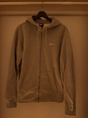Grå kofta från Nike med dragkedja - Grå hoodie från Nike med vit logga på bröstet, hel dragkedja och snörning i huvan. Tröjan har två stora fickor framtill och är tillverkad i mjuk bomullsmix. Perfekt för chill eller träning, klassisk och enkel stil.