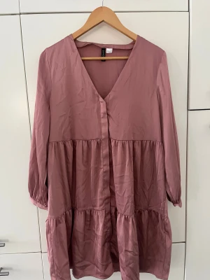 Rosa volangklänning från H&M Divided - Superfin rosa klänning från H&M Divided i storlek 42. Klänningen har v-ringning, långa ärmar med resår vid handlederna och flera volanger som ger en luftig och avslappnad look. Perfekt för dig som gillar en feminin och trendig stil.