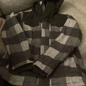 Rutig vinterjacka från Protest, strl 164 - Snygg vinterjacka från Protest i svart och grått rutigt mönster. Jackan har huva, dragkedja och är tillverkad i vattentätt och andningsbart material. Perfekt för kalla dagar och vintersport. Sportig look med coola detaljer och bekväm passform. Använd en vecka. 