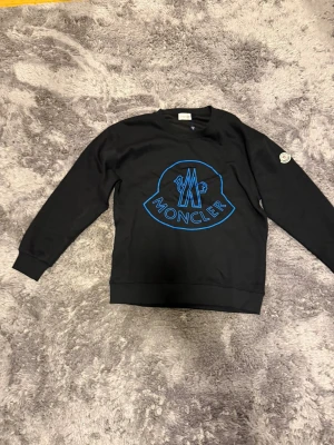 Svart sweatshirt från Moncler med blått tryck - Svart sweatshirt från Moncler med stort blått logotryck på bröstet och klassisk Moncler-patch på ärmen. Tröjan har rund hals, ribbade muddar och är långärmad. Perfekt för dig som gillar streetwear och vill ha något stilrent med tydlig branding.