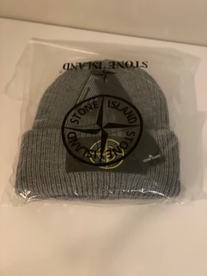 Grå ribbad mössa från Stone Island - Snygg grå mössa från Stone Island med ribbad stickning och klassisk uppvikt kant. Märkets ikoniska kompass-logga sitter framtill och ger en clean streetwear-vibe. Perfekt för dig som vill ha en stilren och varm mössa i vinter.