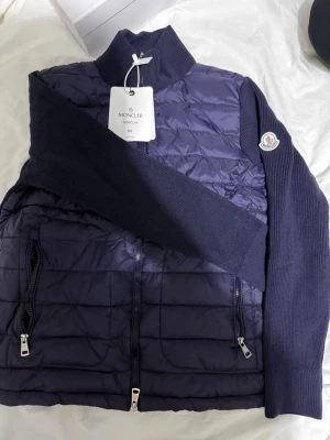 Moncler cardigan  - Snygg mörkblå quiltad jacka från Moncler med stickade ärmar och hög krage. Jackan har två dragkedjefickor framtill, hel dragkedja och klassisk Moncler-logga på ärmen. Perfekt för dig som vill ha en stilren och exklusiv look.