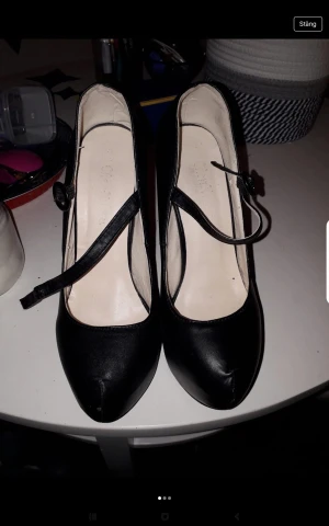 Svarta pumps med hög klack och rem - Säljer ett par svarta pumps med hög klack och rund tå. Skorna har en snygg rem över foten med knappdetalj och plattform framtill. Tillverkade i ett material som liknar skinn och ger en klassisk och stilren look.