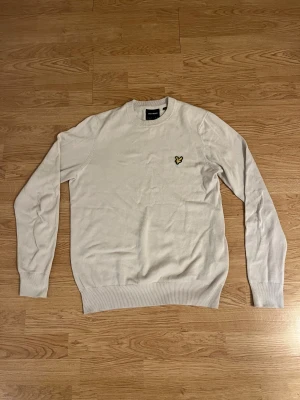 stickad tröja från Lyle & Scott - Begie stickad tröja från Lyle & Scott med klassisk rund hals och långärmad passform. På bröstet sitter den ikoniska gula fågeln som broderad detalj. Tröjan har ribbade muddar vid ärmslut och nederkant, vilket ger en clean och stilren look. Använd 1-2 gånger!! 