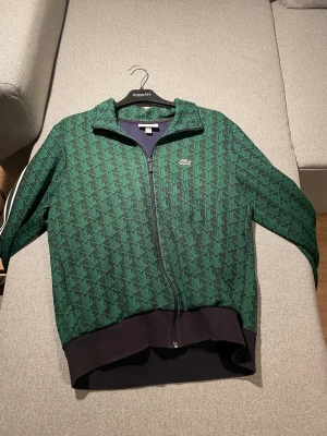 Grön mönstrad track jacket från Lacoste - Snygg grön track jacket från Lacoste med geometriskt mönster i svart och grönt. Jackan har dragkedja framtill, ribbade muddar och vita ränder på axlarna. Regular fit och klassisk Lacoste-logga på bröstet. Perfekt för en sportig och trendig look.