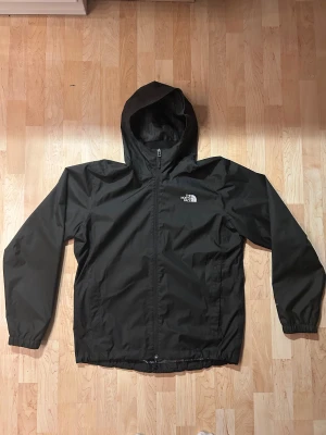 Svart vindjacka från The North Face - Svart vindjacka från The North Face i storlek L. Jackan har huva, hel dragkedja framtill och två sidofickor. Materialet är lätt och syntetiskt, perfekt för blåsiga dagar. Diskret logga på bröstet och elastiska muddar vid ärmslut.