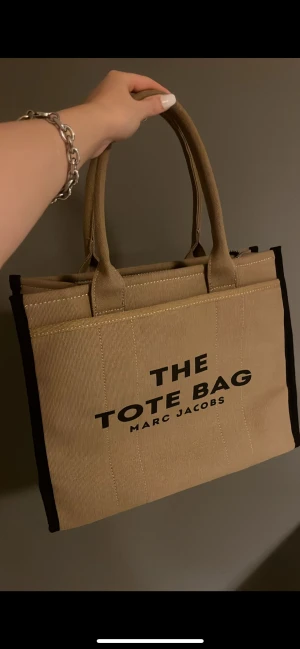 Beige tote bag från Marc Jacobs - Snygg och rymlig tote bag från Marc Jacobs i beige canvas med svarta detaljer och tydlig logga framtill. Väskan har två stadiga handtag och en klassisk fyrkantig form, perfekt för att bära med sig allt man behöver.