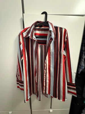 Randig skjorta från Gina Tricot över size 🔴 - Snygg skjorta från Gina Tricot i storlek S med vertikala ränder i rött, ljusblått, vitt och mörkbrunt. Skjortan har klassisk krage, lång ärm med knapp vid manschetten och en bröstficka. Materialet är mjuk bomull som känns skönt mot huden.