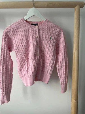 Rosa kofta från Ralph Lauren - Snygg rosa kofta från Ralph Lauren i barnstorlek L. Vuxen storlek S. Den är kabelstickad i 100% bomull och har knappar framtill samt det klassiska gröna Ralph Lauren-logot på bröstet. Rund hals och långa ärmar ger en chill och preppy vibe.