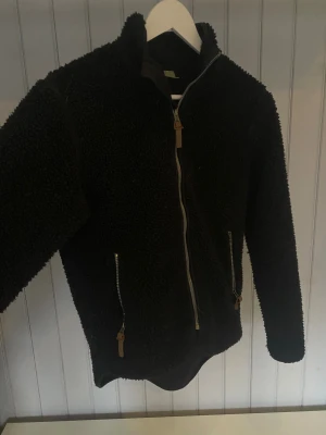 Svart teddyjacka med dragkedja - Mysig svart teddyjacka med hög krage och hel dragkedja framtill. Jackan har två sidofickor med dragkedjor och detaljer i läderimitation. Använd fåtal gånger. I storlek 36/38