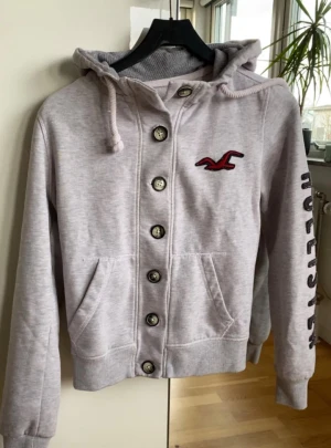 Ljusgrå hoodie från Hollister med knappar - Ljusgrå hoodie från Hollister med knappar framtill, broderad röd logga på bröstet och svart Hollister-text på ärmen. Tröjan har huva med snörning, två fickor och ribbade muddar. Perfekt för chill dagar och snygg streetstil.
