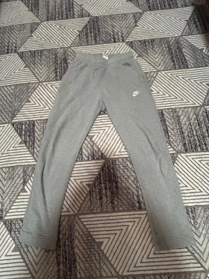 Grå mjukisbyxor från Nike, storlek S - Grå mjukisbyxor från Nike med klassisk logga broderad på vänster lår. Byxorna har elastisk midja och raka ben, tillverkade i mjukt bomullsmaterial som är skönt mot huden. Perfekta för chill eller träning, med fickor på sidorna.