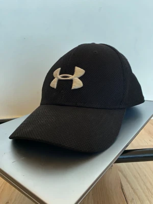 Svart keps från Under Armour -stolek one size - Svart keps från Under Armour med böjd skärm och vit broderad logga framtill. Baksidan har Under Armour-text i vitt. Tillverkad i ventilerande meshmaterial, 97% polyester och 3% spandex. Perfekt för sport eller chill dagar.