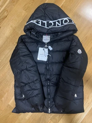  Moncler  - Svart dunjacka från Moncler med stor huva och vit logotyptext på kanten. Jackan har quiltad design, dragkedja framtill och Moncler-märke på ärmen. Insidan har en cool serietidningsetikett. Perfekt för dig som vill ha en snygg och varm jacka med street vibe.                      Passar M/L